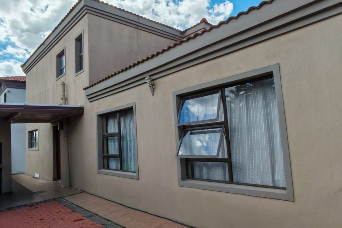Rear view of the property featuring a plastered facade, aluminium window frames, and a covered patio area with paved flooring.