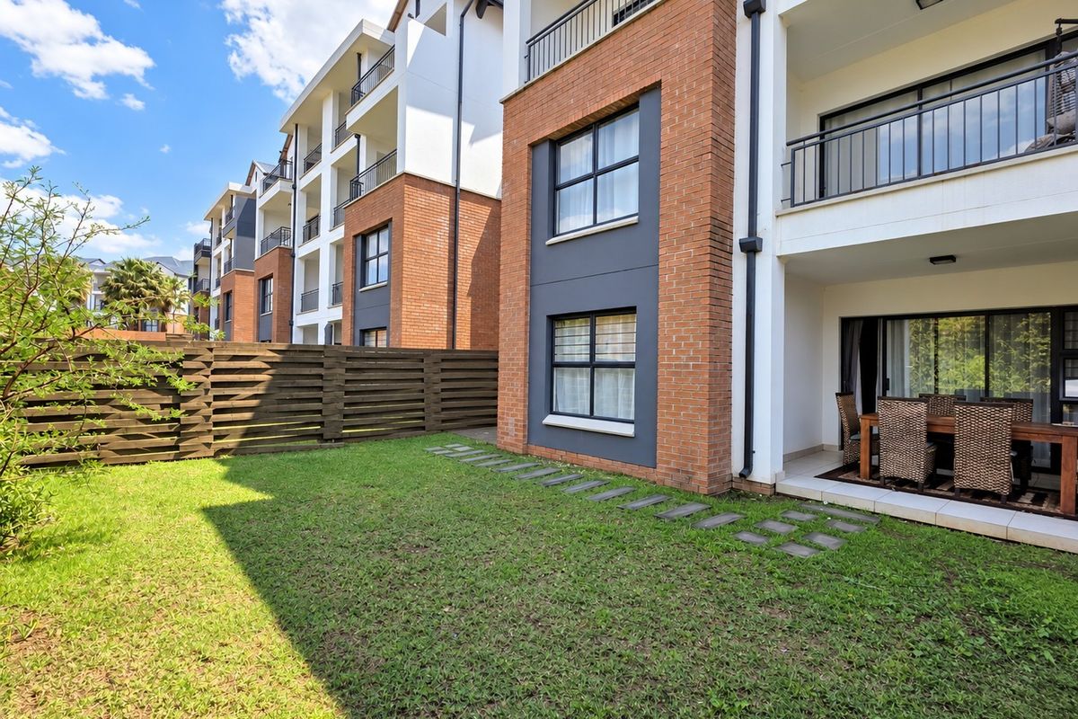 A ground floor apartment exterior featuring a private garden, paved patio area, and brick facade with balcony access above.
