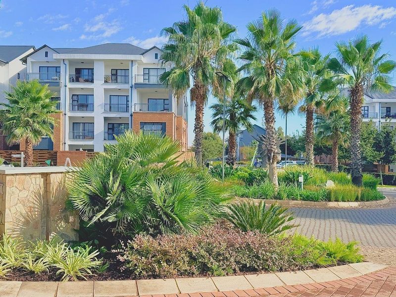 A well-maintained apartment complex entrance featuring landscaped gardens, paved driveways, and modern multi-story residential buildings with private balconies.