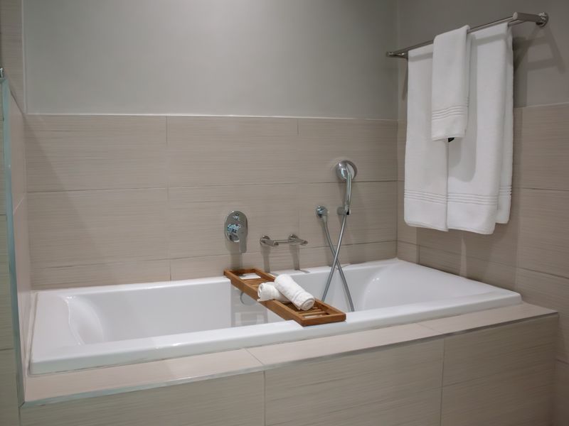 A modern bathroom featuring a built-in bathtub with wall-mounted chrome fixtures, handheld shower attachment, and neutral-toned tiled wall finishes.