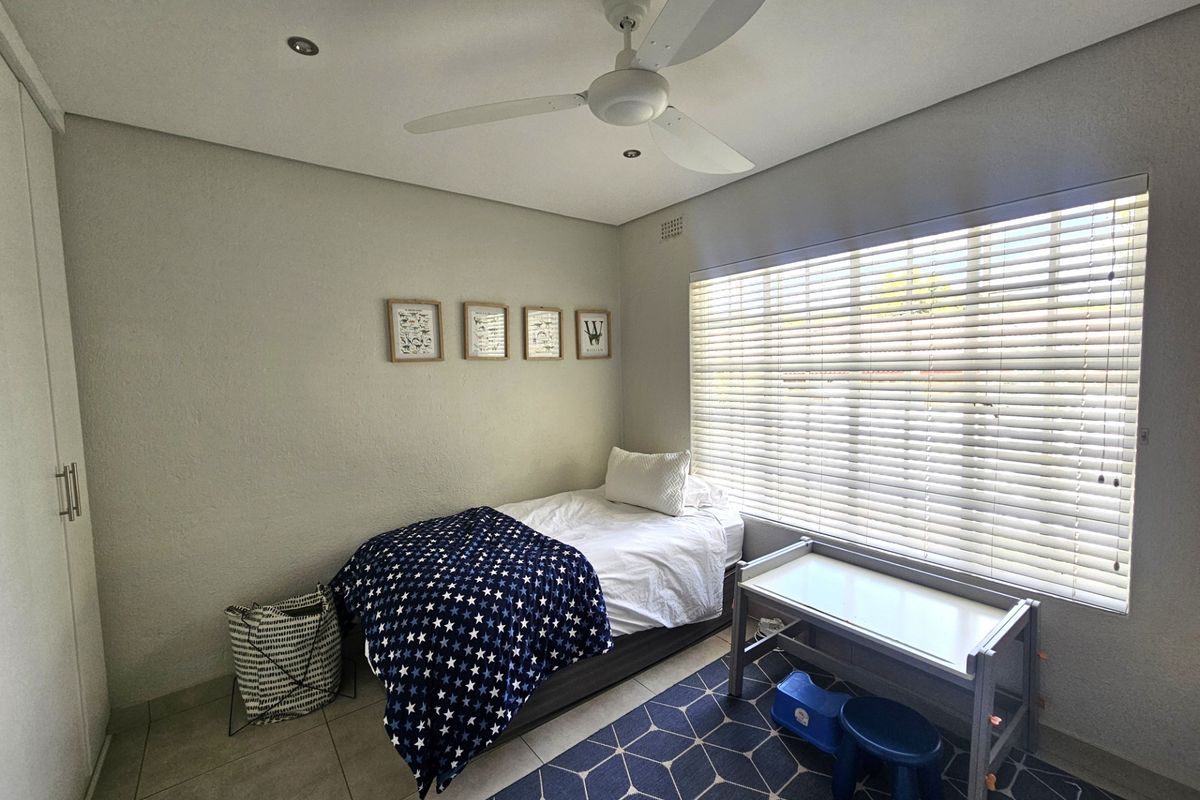A well-maintained bedroom featuring tiled flooring, built-in cupboards, a ceiling fan, and large windows fitted with venetian blinds.