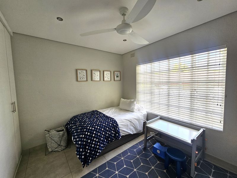 A well-maintained bedroom featuring tiled flooring, built-in cupboards, a ceiling fan, and large windows fitted with venetian blinds.