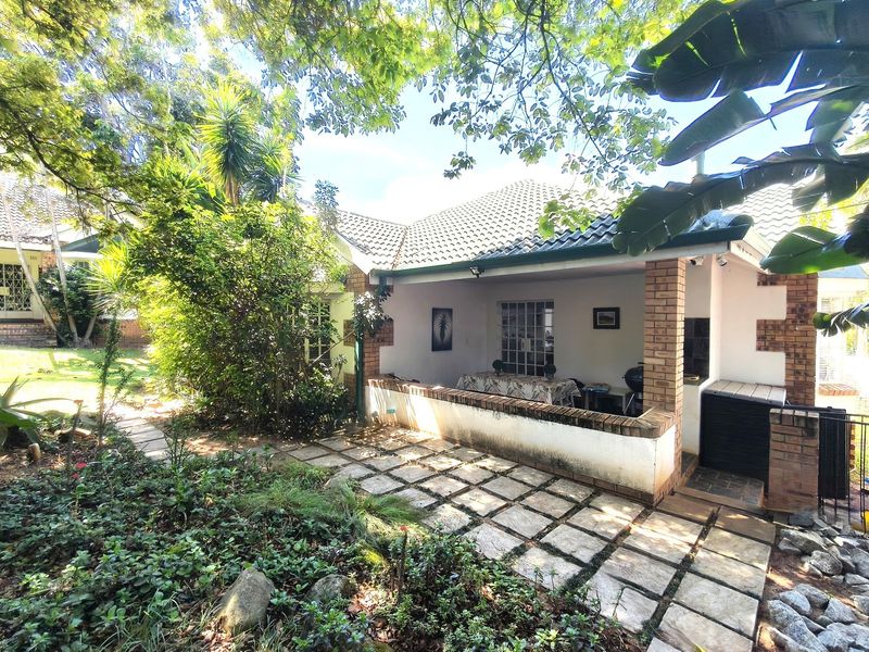 Covered patio area featuring brick pillars, built-in braai, and paved flooring, overlooking a lush garden setting.