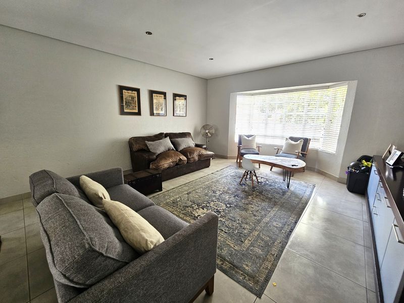 A bright and spacious living room featuring large bay windows with venetian blinds, recessed downlighting, and neutral tiled flooring.