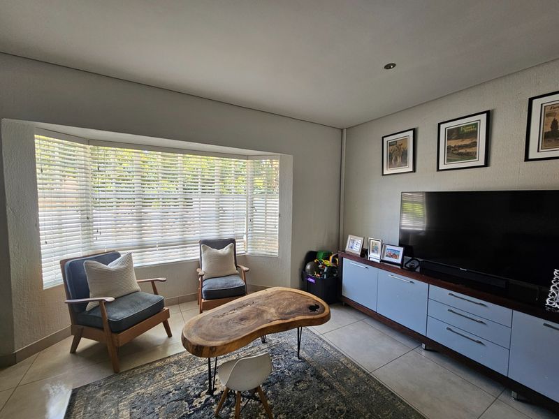 A bright living room featuring large bay windows with venetian blinds, neutral tiled flooring, and recessed downlighting, creating a comfortable and well-lit space.