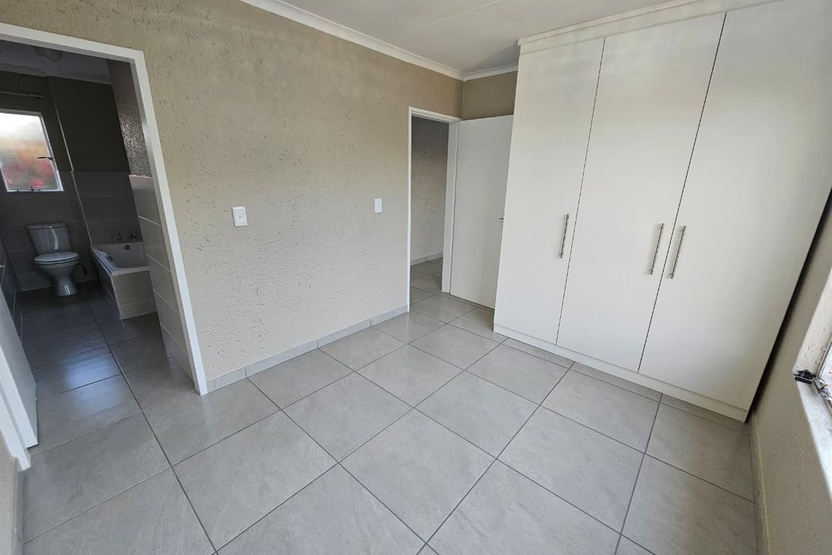 A clean, empty bedroom featuring neutral tiled flooring, built-in white cupboards, and direct access to an en-suite bathroom.