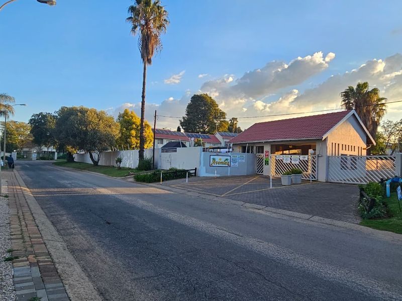Street view of the Avonlea townhouse complex entrance, featuring a paved driveway, secure gated access, and perimeter walling with electric fencing.