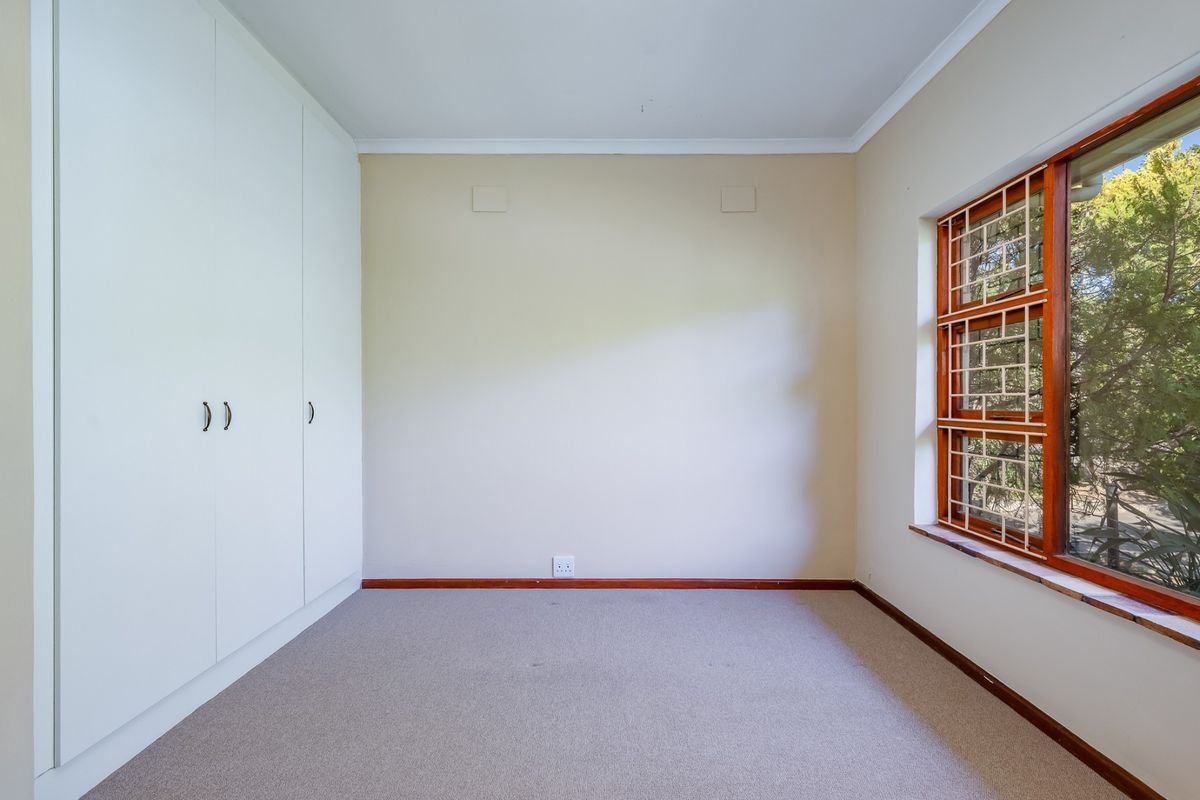 A clean, empty bedroom featuring neutral walls, carpeted flooring, and ample built-in cupboard storage with a window overlooking the garden.