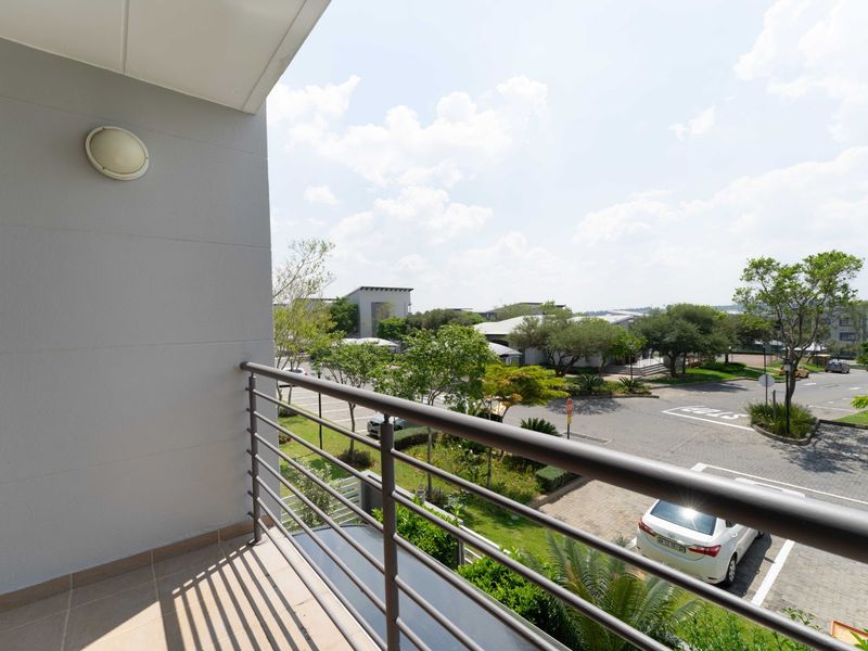 A clean, tiled balcony featuring a metal safety railing and an exterior wall light, overlooking the complex grounds and parking area.