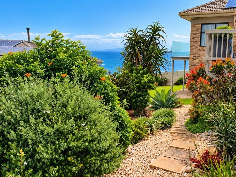 A well-maintained rear garden featuring a stone path, lush landscaping, and a timber deck with glass balustrades, offering scenic ocean views.