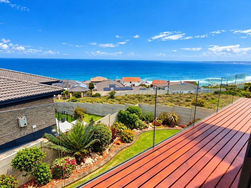 Elevated view from a timber deck featuring a glass balustrade, overlooking a landscaped garden, residential neighborhood, and the ocean.