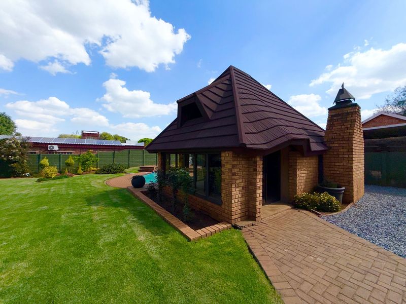 A well-maintained rear garden featuring a brick outbuilding with a chimney, a paved walkway, and a swimming pool set within a landscaped lawn.