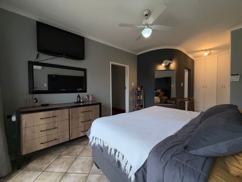 A spacious bedroom featuring tiled flooring, built-in cupboards, and a ceiling fan, with a neutral grey color scheme.