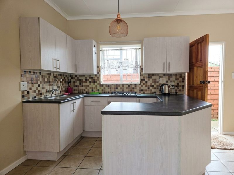 A functional kitchen featuring light wood-grain cabinetry, dark countertops, mosaic tile backsplash, and a built-in gas hob with direct access to the exterior.