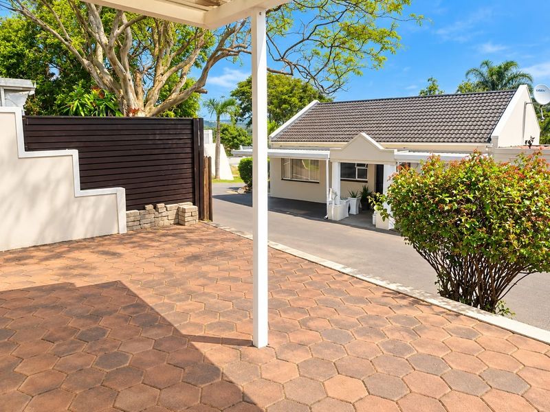 A paved outdoor patio area featuring a covered roof structure and timber privacy screen, overlooking the complex driveway and surrounding garden.