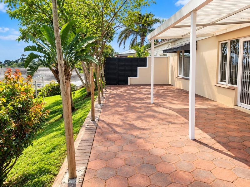 A spacious paved patio area featuring a pergola, overlooking a well-maintained garden with established trees and secure boundary walls.