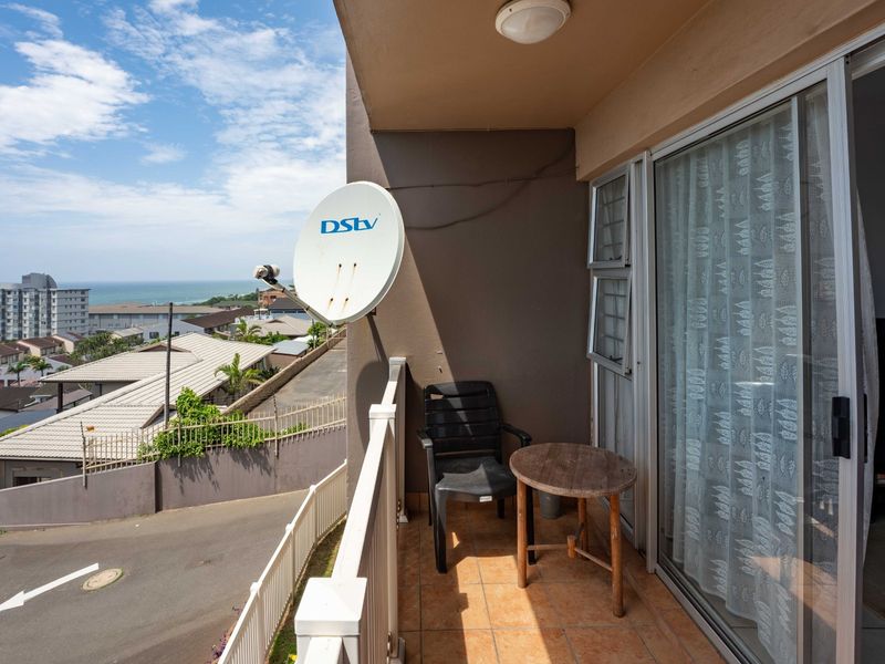 A covered balcony featuring tiled flooring, a metal railing, and sliding glass door access, offering elevated views of the surrounding neighborhood and coastline.