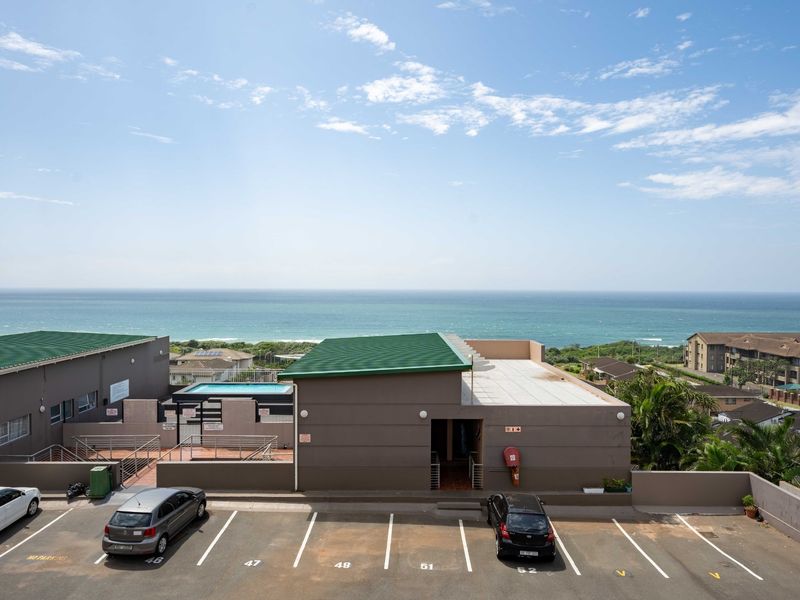 Exterior view of the apartment complex featuring a paved parking area, green metal roofing, and a swimming pool with ocean views.