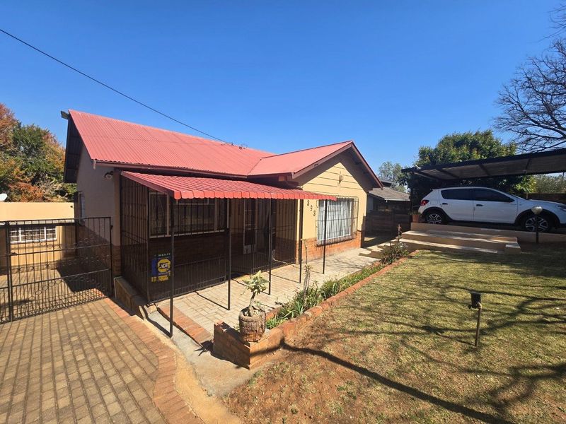 Front elevation of a single-story house featuring a red corrugated metal roof, paved driveway, security gates, and a carport.