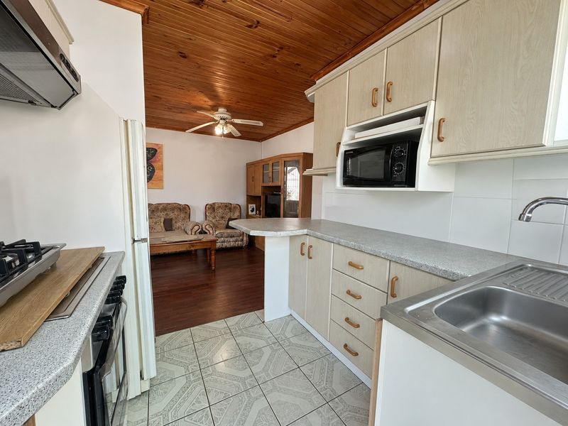 Open-plan kitchen featuring light-toned cabinetry, laminate countertops, and integrated appliances, flowing into a living area with timber ceilings and tiled flooring.