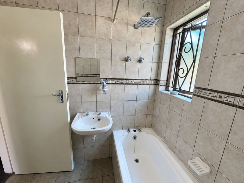 A functional bathroom featuring a built-in bathtub, wall-mounted basin, and overhead shower, finished with neutral wall tiles and a secure window.
