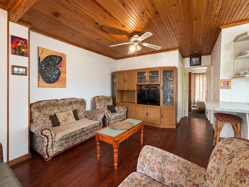 A cozy living room featuring warm timber ceilings, hardwood flooring, and a built-in entertainment unit, with an open flow toward the kitchen and hallway.