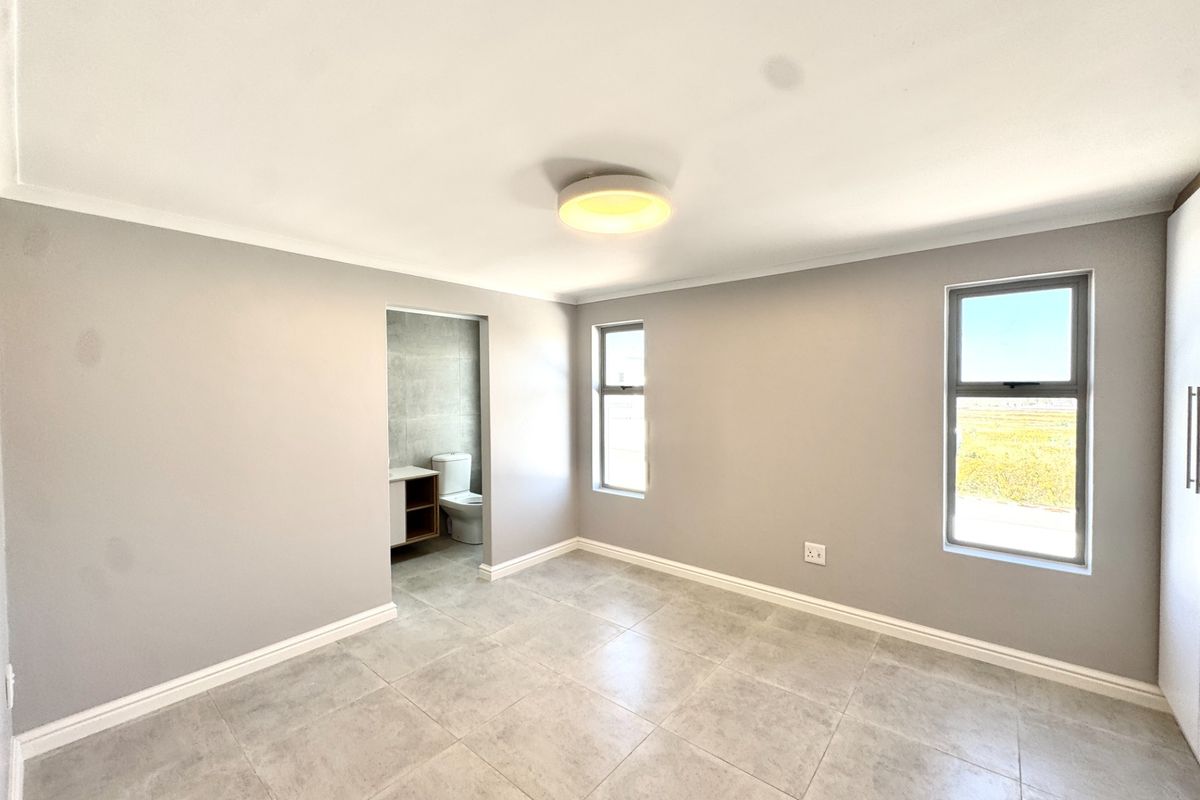 A bright, modern bedroom featuring neutral grey walls, tiled flooring, built-in cupboards, and an en-suite bathroom access.