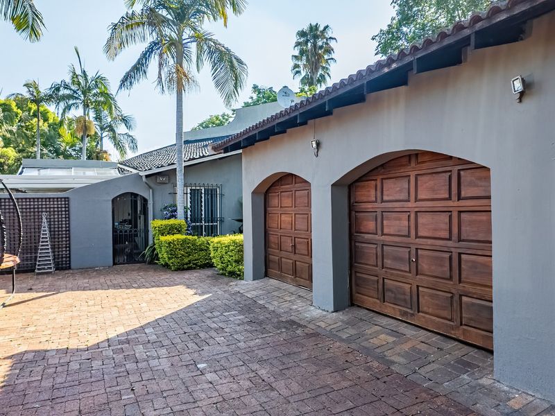 Front exterior view featuring a double garage with wooden doors, brick paved driveway, and established garden surroundings.