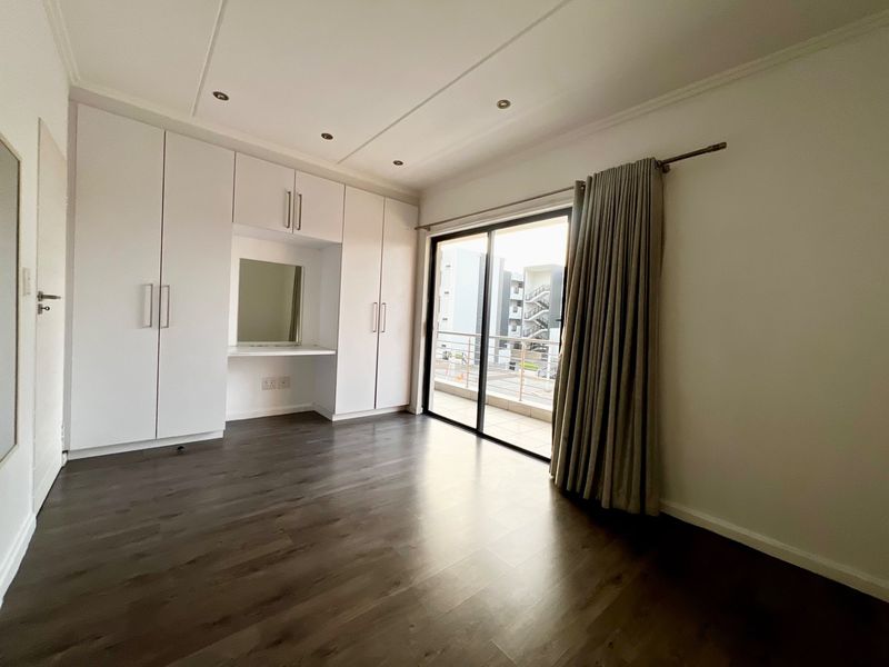 A bright, empty bedroom featuring modern laminate flooring, extensive built-in cupboards with an integrated vanity, and sliding door access to a private balcony.