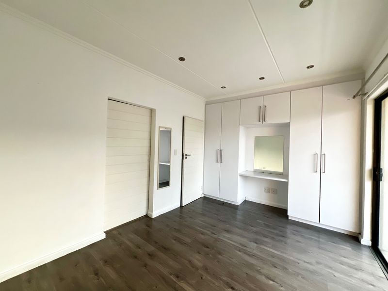 A bright, empty bedroom featuring modern laminate flooring, recessed downlights, and extensive built-in cupboard storage with an integrated vanity area.