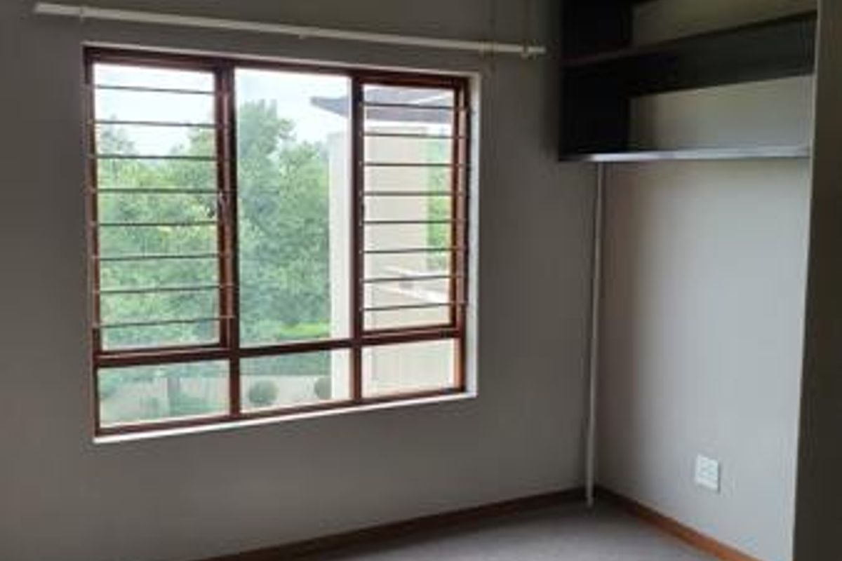 A bright, empty bedroom featuring neutral carpeting, a ceiling fan, and built-in wall shelving, with large windows fitted with security bars.