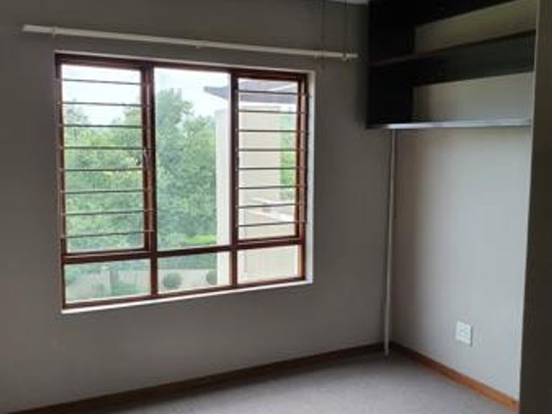 A bright, empty bedroom featuring neutral carpeting, a ceiling fan, and built-in wall shelving, with large windows fitted with security bars.