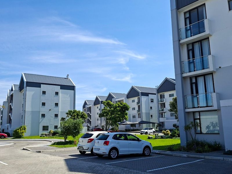 Exterior view of a modern apartment complex featuring multi-story buildings with pitched roofs, glass-fronted balconies, and a paved parking area.