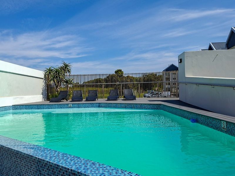 A well-maintained outdoor swimming pool area featuring blue mosaic tiling, surrounding decking, and secure perimeter fencing under a clear blue sky.