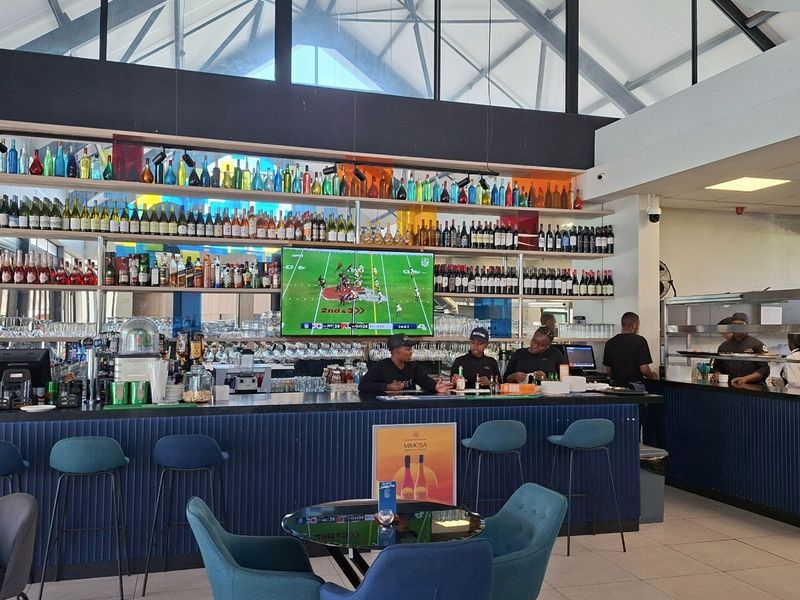 A modern, well-maintained bar area featuring a long built-in counter, extensive back-bar shelving, and a high vaulted ceiling with large windows.