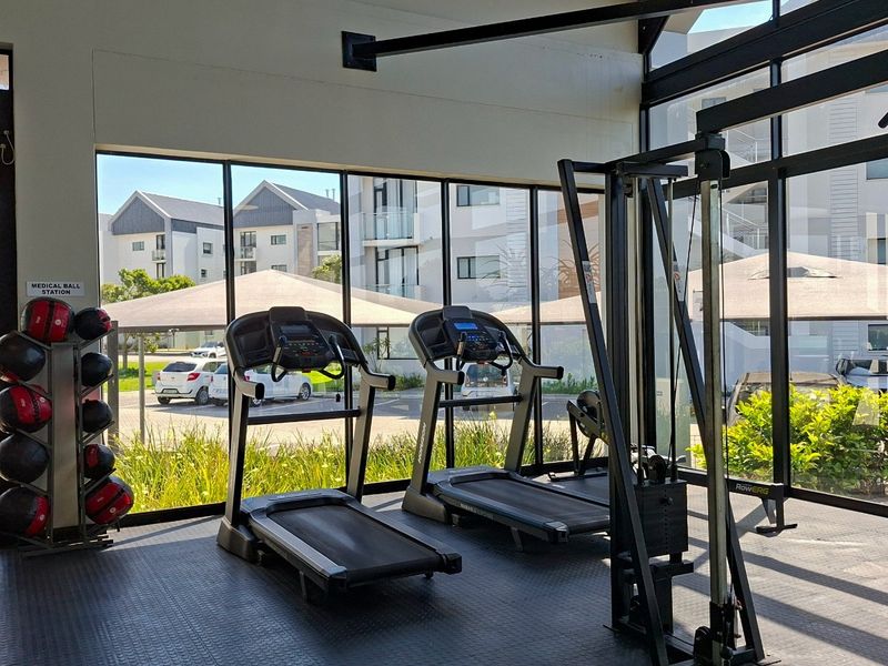 A modern, well-lit gym facility featuring durable rubber flooring, floor-to-ceiling windows, and views of the surrounding apartment complex.