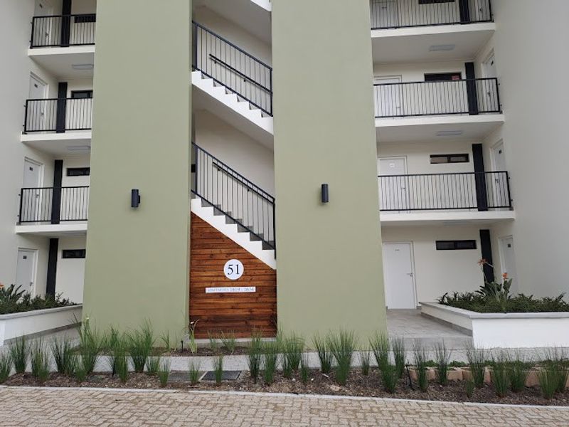 Modern apartment complex exterior featuring a central staircase, clean facade, metal railings, and well-maintained landscaped garden beds at the entrance.