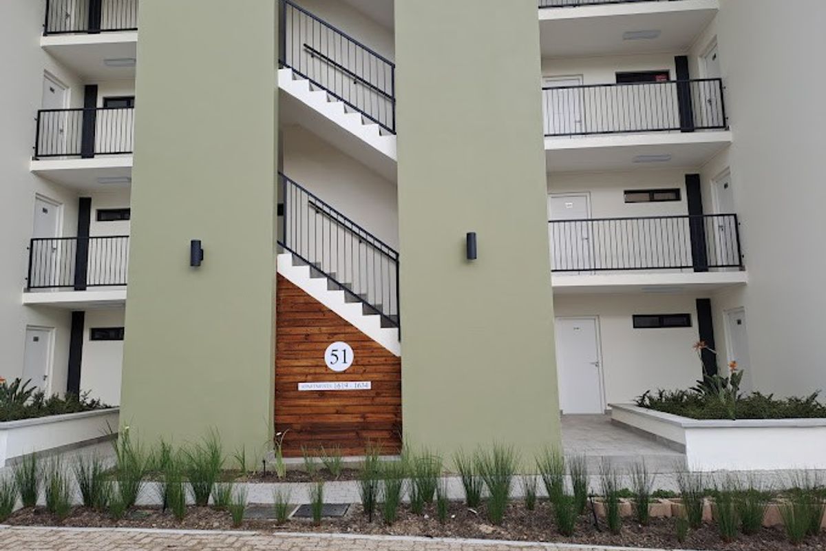 Modern apartment complex exterior featuring a central staircase, clean facade, metal railings, and well-maintained landscaped garden beds at the entrance.