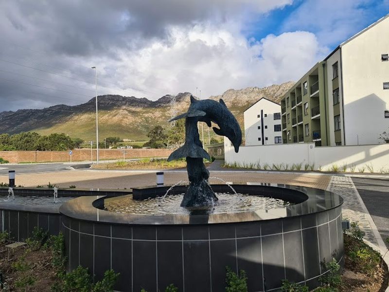 A well-maintained apartment complex entrance featuring a decorative dolphin water fountain, paved driveway, and scenic mountain views.