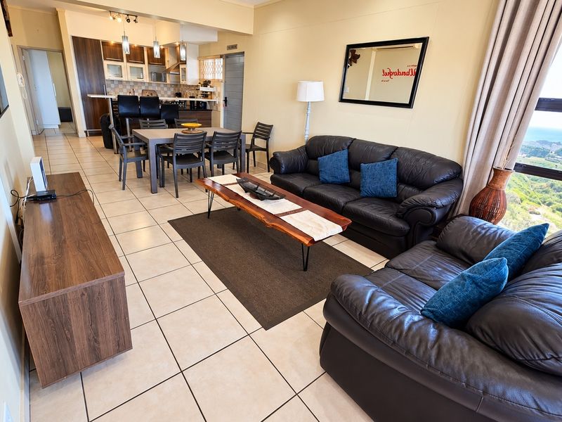 Spacious open-plan living and dining area featuring tiled floors, a modern kitchen with breakfast bar, and large windows offering scenic coastal views.