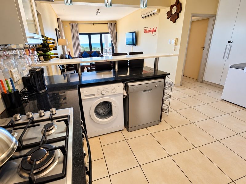 Open-plan kitchen and living area featuring granite countertops, a gas hob, tiled flooring, and integrated storage, leading to a balcony with ocean views.