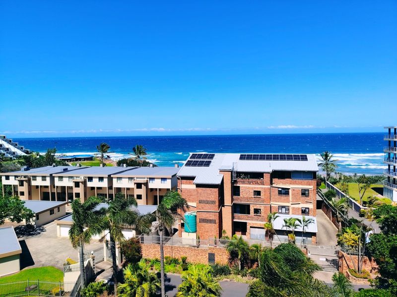 An aerial view of a multi-story brick apartment complex featuring a metal roof with solar panels, situated near the coastline.