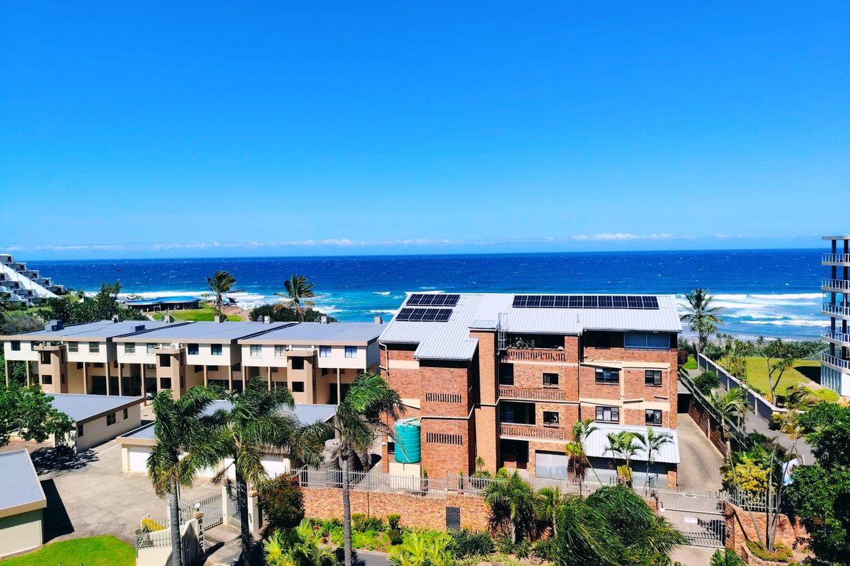 An aerial view of a multi-story brick apartment complex featuring a metal roof with solar panels, situated near the coastline.