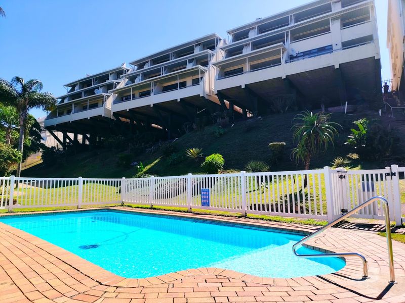 A well-maintained communal swimming pool area featuring brick paving, safety fencing, and a stainless steel ladder, set against a multi-level apartment complex.