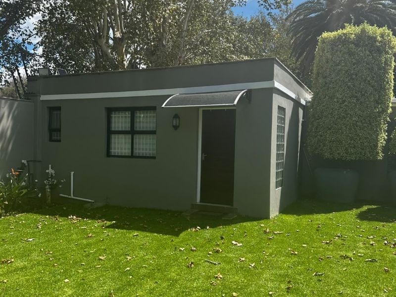 A well-maintained detached cottage structure featuring a modern grey facade, glass block accents, and a small entrance awning set within a garden.