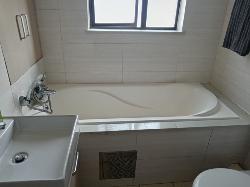 A functional bathroom featuring a built-in bathtub with shower mixer, wall-mounted basin, and neutral tiled finishes throughout.