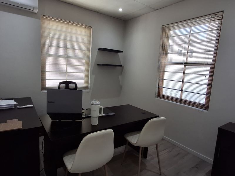 A functional office space featuring laminate flooring, dual windows with venetian blinds, floating shelving, and a wall-mounted air conditioning unit.