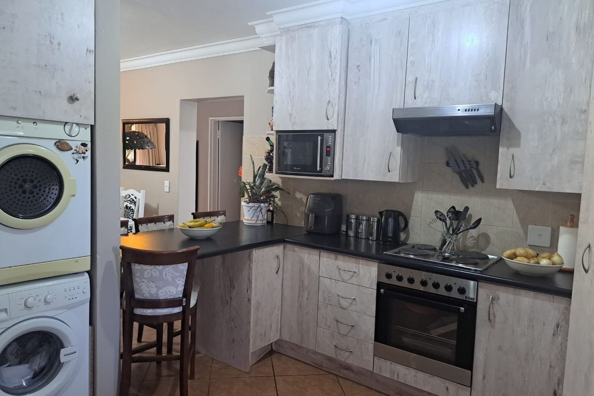 A functional kitchen featuring light wood-grain cabinetry, dark countertops, built-in oven and hob, and a dedicated laundry area with stacked appliances.