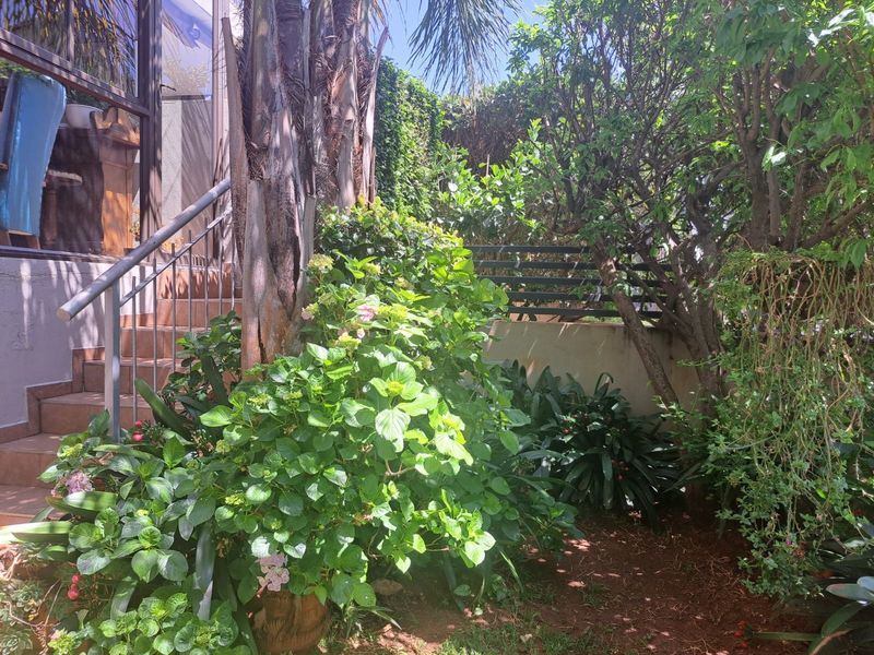 A lush garden area featuring mature trees, established shrubs, and a tiled staircase with a metal handrail leading to the property entrance.