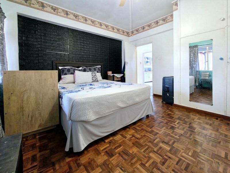A bedroom featuring classic parquet flooring, built-in cupboards with a mirror, and a ceiling fan, offering a functional space with potential for modernization.